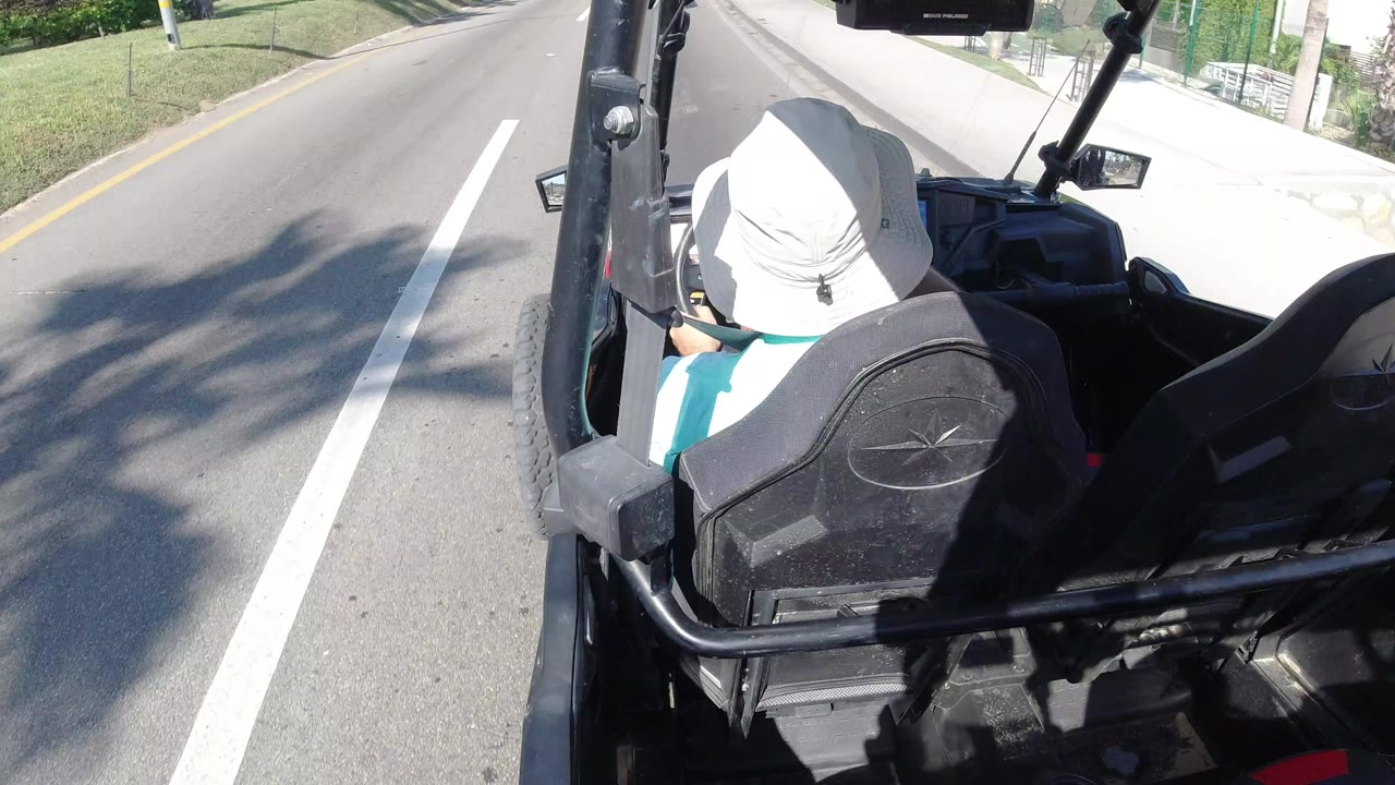 Rear-mounted view of a driver in a white hat inside an open off-road vehicle on a bright Los Cabos street, with lane markings and a grassy roadside visible.