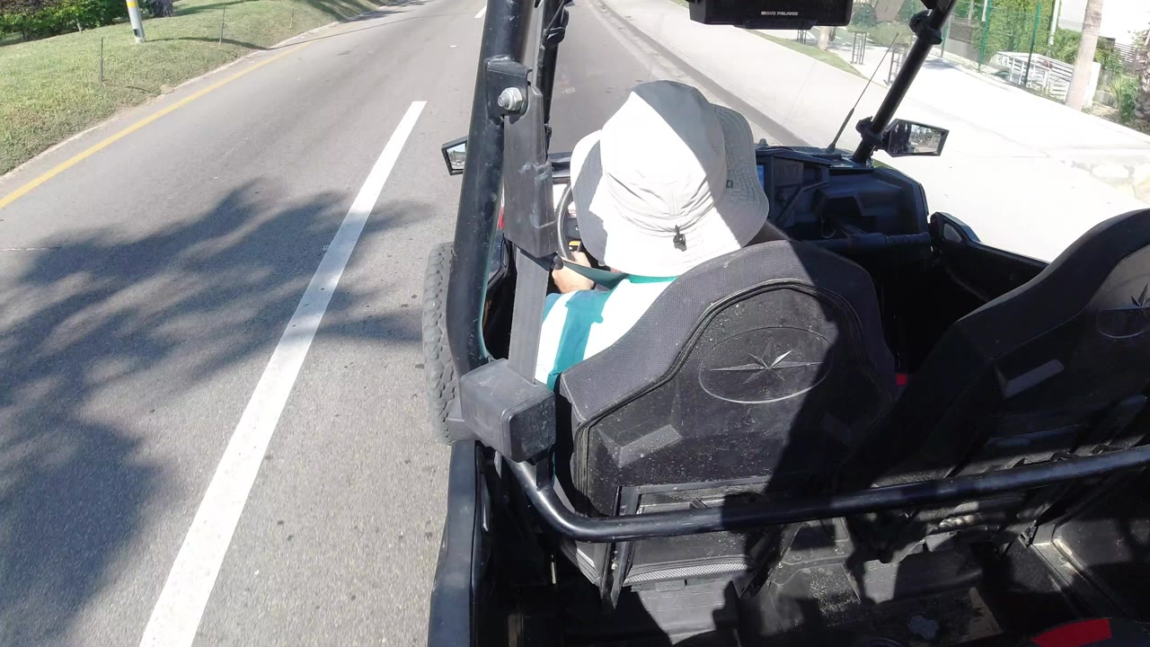 The driver remains framed from behind in the open vehicle as the Los Cabos street and curb slide past under strong sunlight.