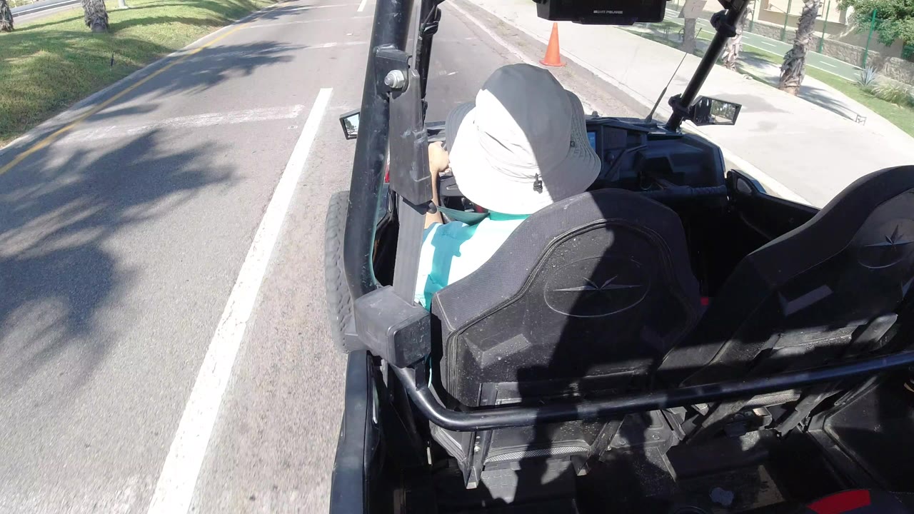 Rear view of a man in a light brimmed hat driving an open jeep along a bright paved road in Los Cabos, Mexico, with lane markings and roadside grass visible.