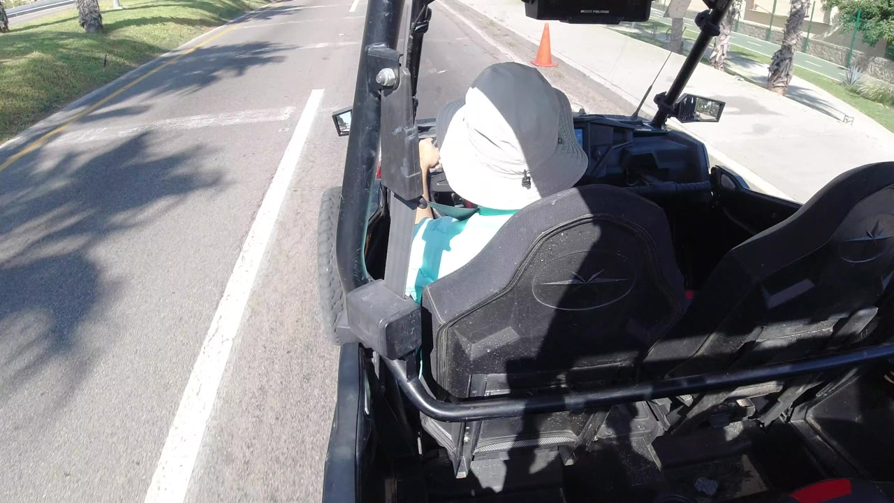 The Los Cabos road view continues from behind the driver, with the black jeep seat backs in the foreground and a sidewalk, curb, and orange cone ahead.