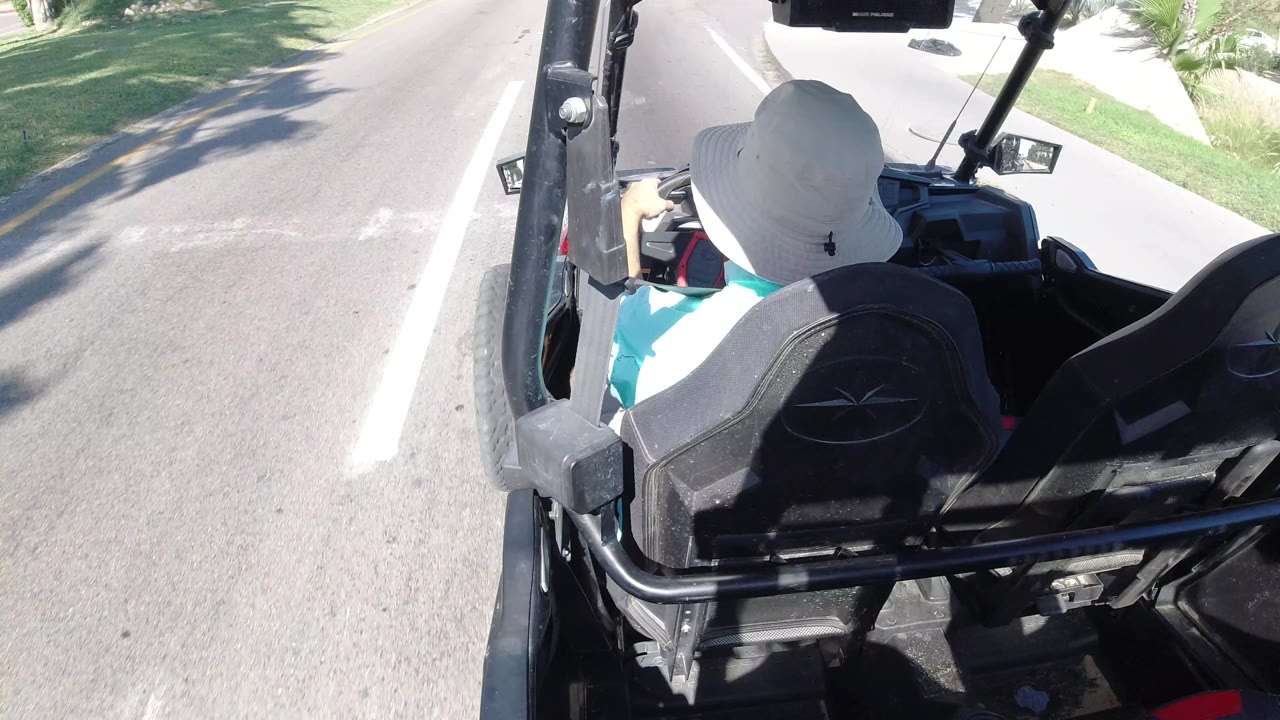 At 0:01, the camera remains high behind the buggy rider, showing black seats, metal supports, road surface, a grassy verge, and bright sunlight in Los Cabos, Mexico.