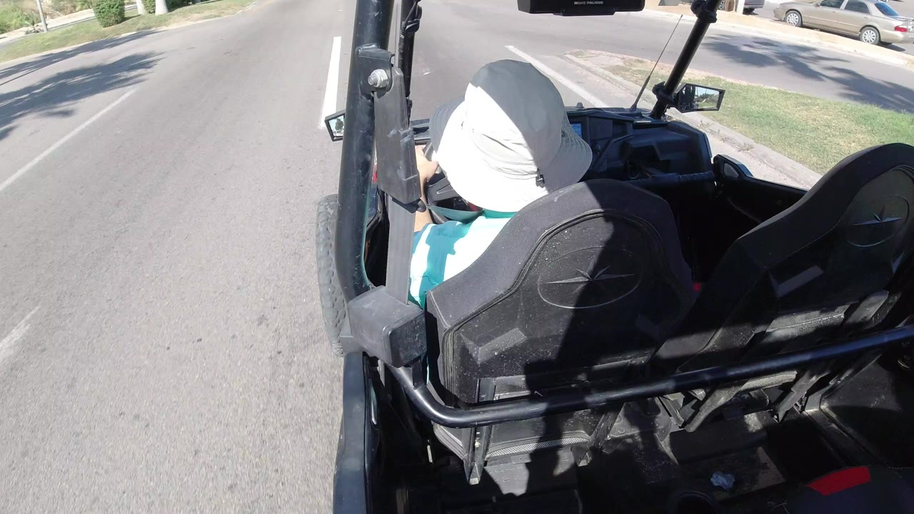 Open vehicle moving along a sunlit street with a seated passenger in a white hat.