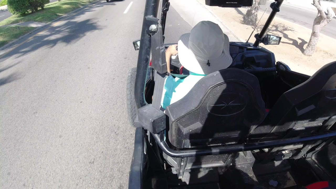 The Los Cabos road view shows the driver from behind as a white car appears beside the open vehicle on the sunlit street.