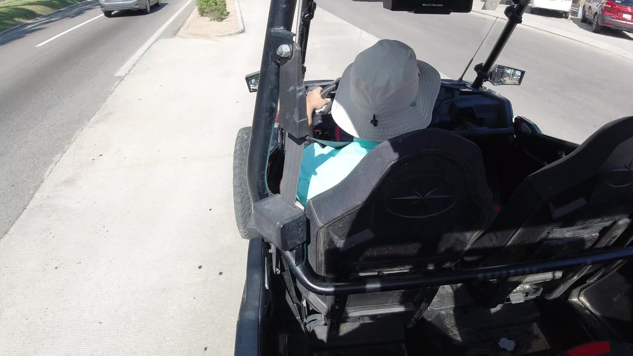 Rear view from above a black dune buggy driving on a Los Cabos street, with the hat-wearing driver seen from behind, curb, sidewalk, and passing cars visible.