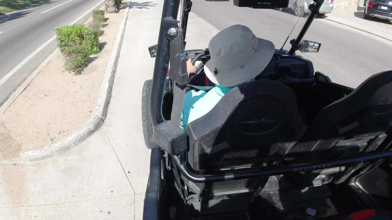 The dune buggy continues along a Los Cabos road with the driver viewed from behind, a sandy median or planting strip, low shrubs, sidewalk, and parked cars around the street.