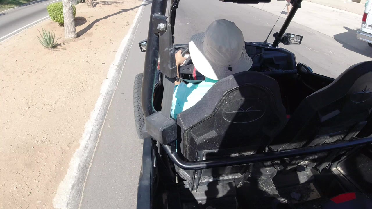 Rear high-angle view in Los Cabos of an open vehicle with black seats, a driver in a white hat, sandy landscaping, curb, and paved road visible.