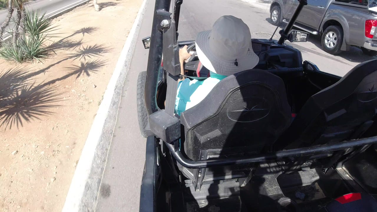 A rear overhead view follows the hat-wearing driver in the open buggy as a gray pickup appears along the street beside dry landscaping in Los Cabos, Mexico.