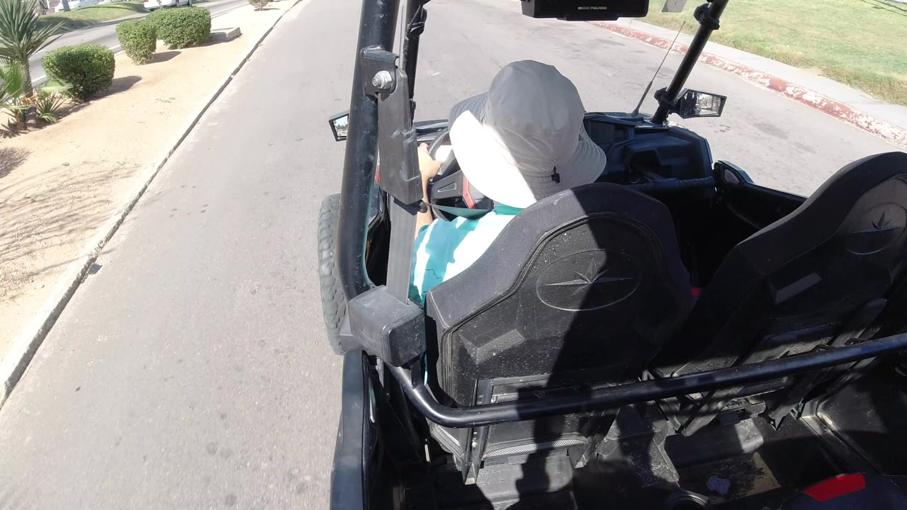 A person in a white hat sits at the front of a black golf cart in Los Cabos, Mexico, while the road and curb run past landscaped shrubs in bright sun.