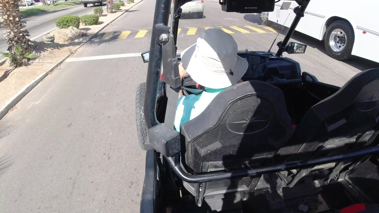 The open vehicle continues along the Los Cabos road, with the driver seen from behind, black seats and roll bars in the foreground, a white bus to the right, and palms and shrubs along the divider.