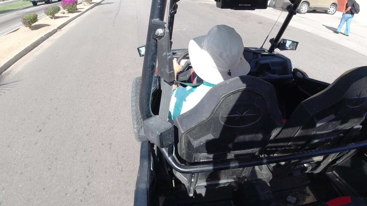 In Los Cabos, Mexico, the camera stays behind the white-hatted driver as the open vehicle continues along a mostly clear paved road with sidewalk planting and parked cars near the curb.