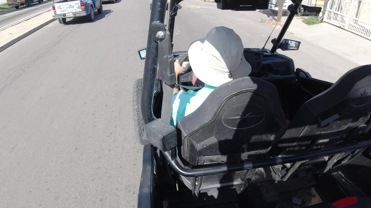 At 0:01 in Los Cabos, Mexico, the driver remains centered from behind as the open vehicle follows the paved street past parked cars and low residential frontage.