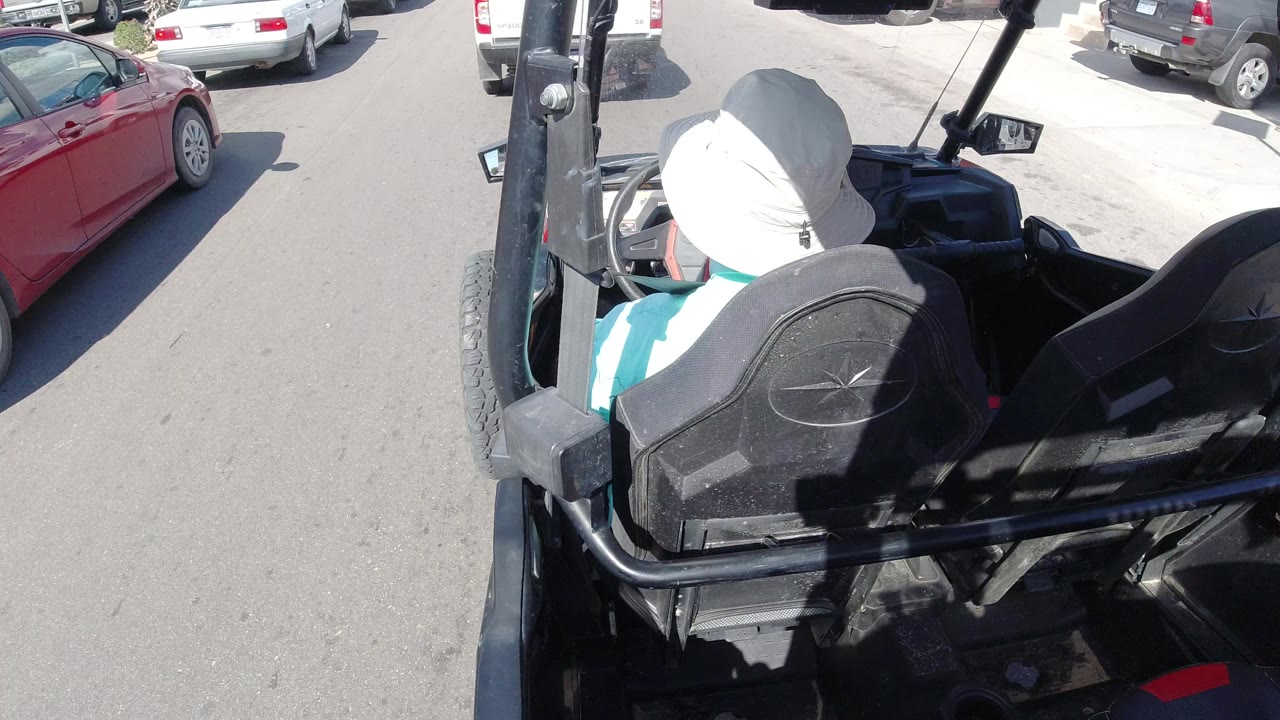 A person in a white hat sits in a black open vehicle on a sunny Los Cabos street, with a red car and white vehicles visible nearby.