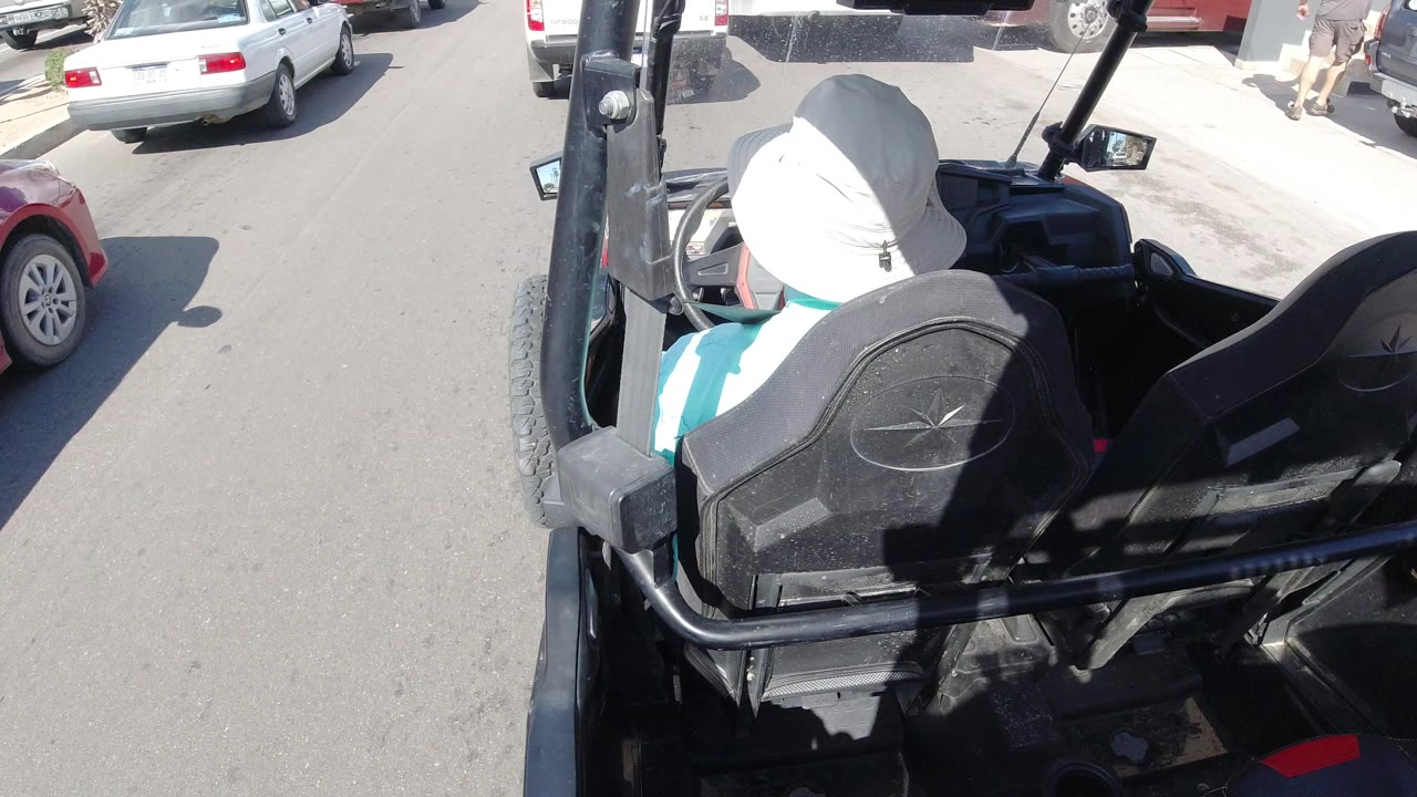 Man in a white hat seated in an open black vehicle on a sunny street.