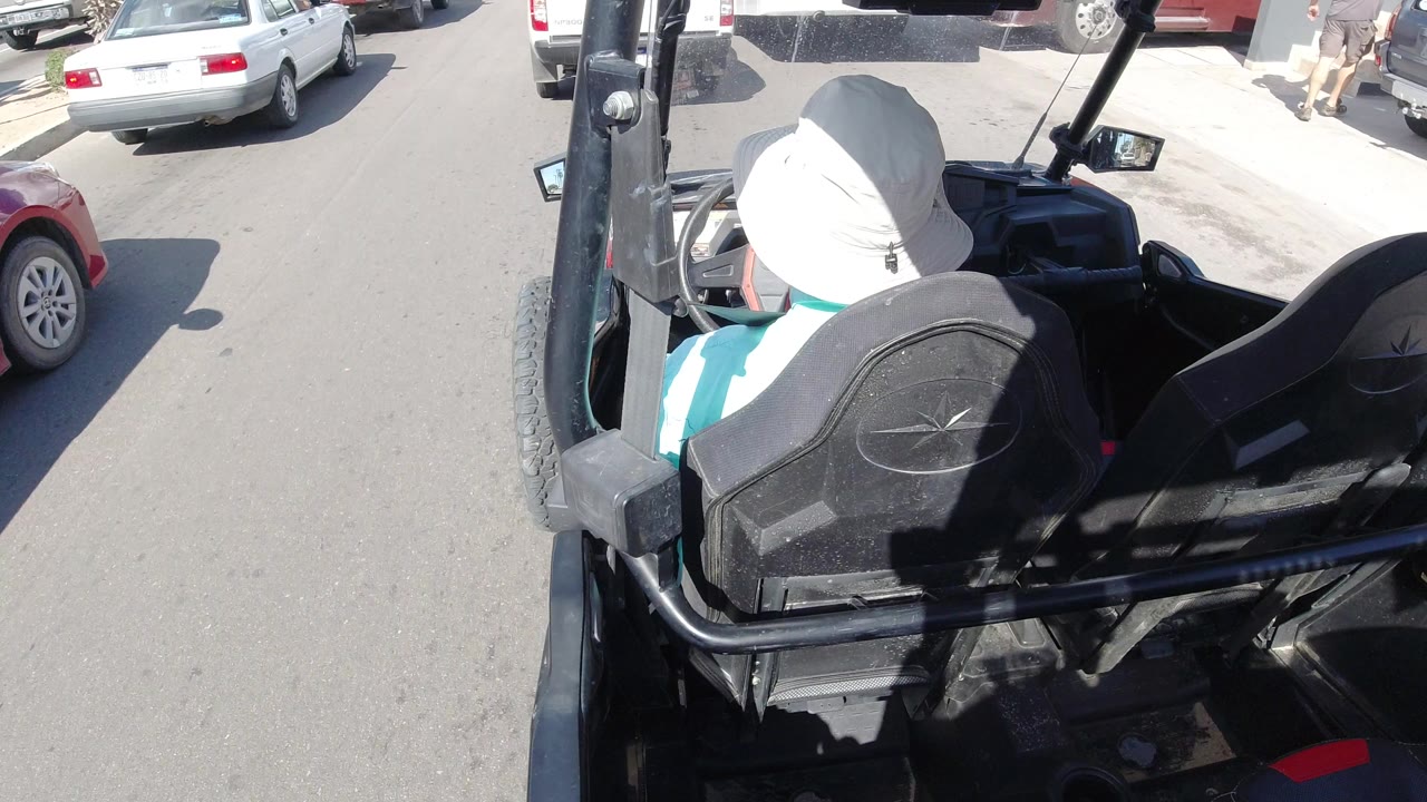 Rear elevated frame of the white-hatted driver in the open vehicle, with the street and a white parked car visible beyond the front corner.
