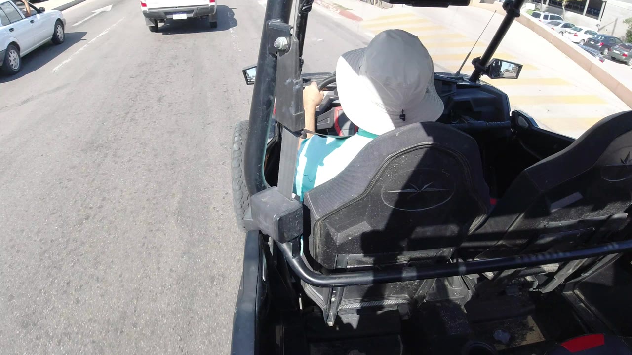 The open vehicle follows traffic in Los Cabos, Mexico, with the driver's white hat centered, a white pickup ahead, parked cars along the curb, and strong sunlight on the asphalt.