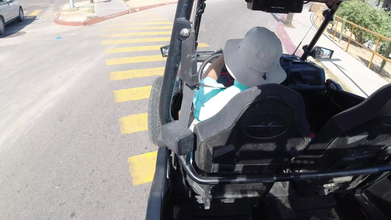 In Los Cabos, Mexico, a woman seen from behind drives an open black mini vehicle across yellow crosswalk markings on a bright paved road.