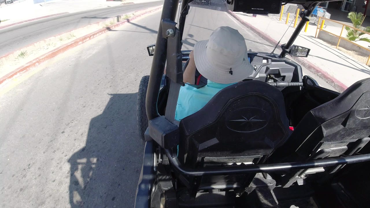 The driver remains centered from behind as the open cart approaches a sidewalk and building frontage on a bright Los Cabos street.