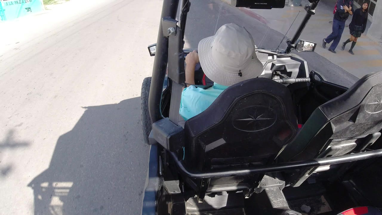 Opening frame from Los Cabos, Mexico shows the back of a black golf cart, a driver in a pale hat, bright pavement, and distant pedestrians near the upper right.