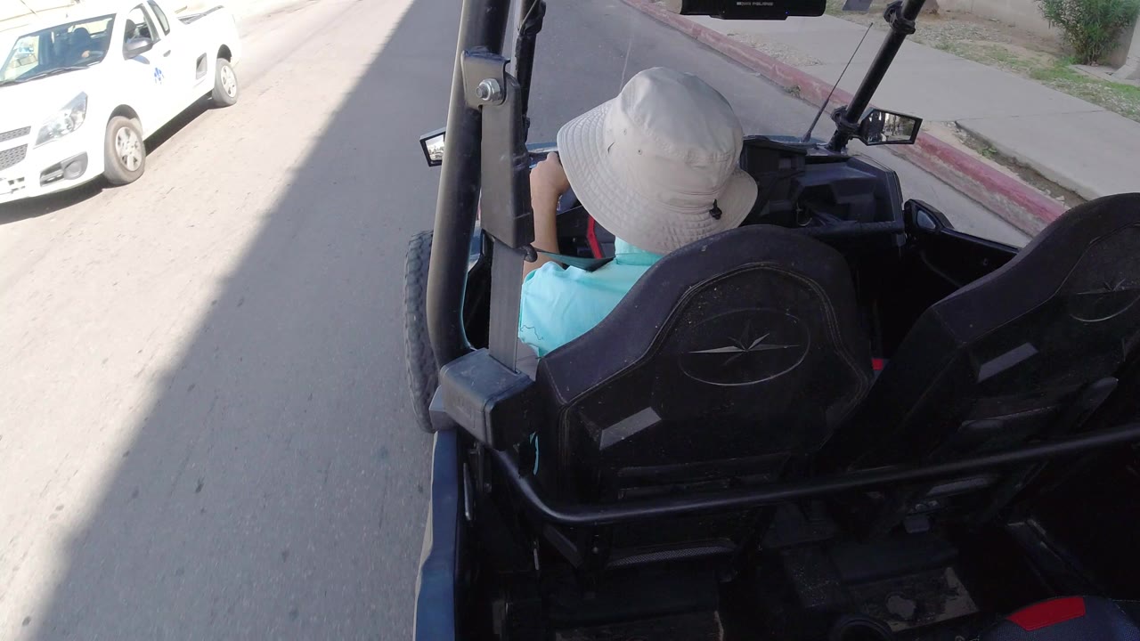 Rear view in Los Cabos, Mexico of a person in a white sun hat and turquoise shirt seated in a black golf cart beside a bright sandy road and a white car.