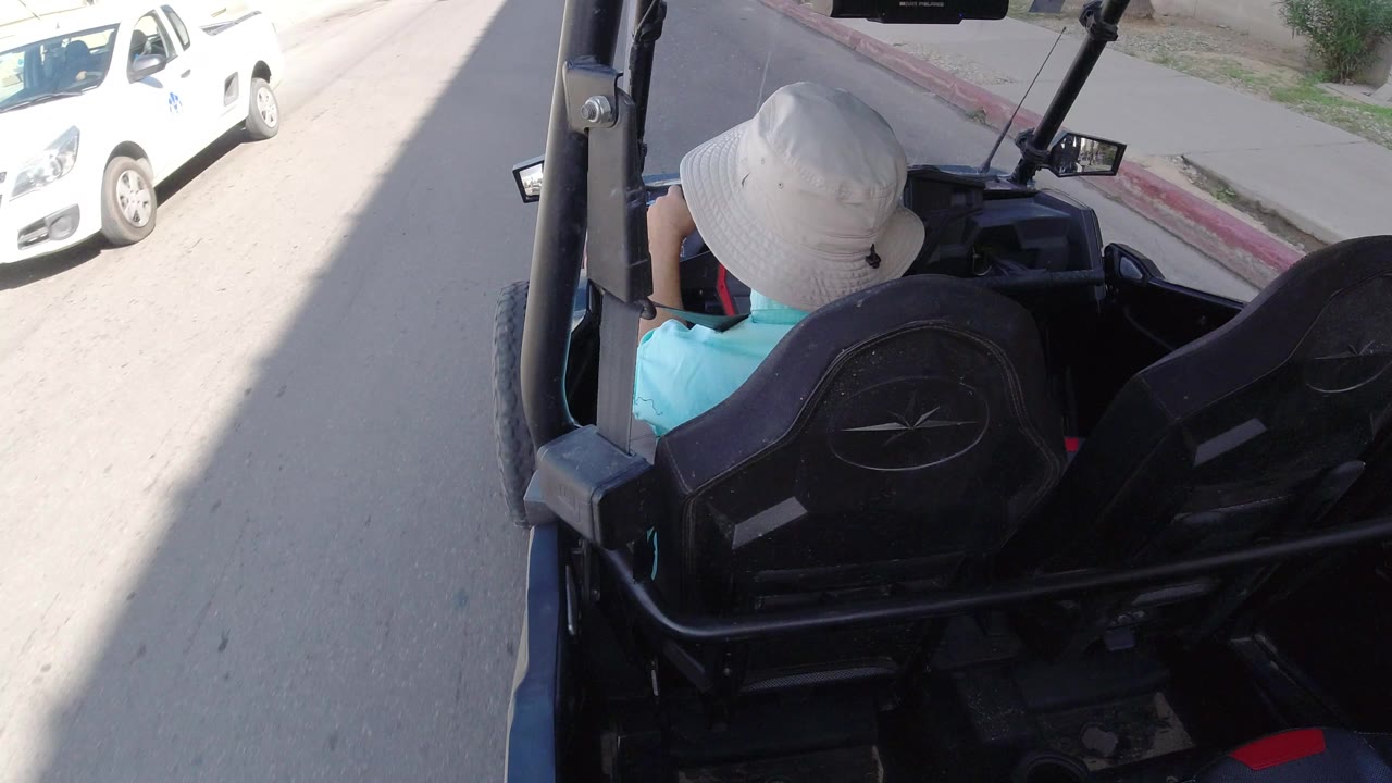 In Los Cabos, Mexico, the white-hatted passenger remains framed from behind in the black golf cart as the pale road and red curb run along the right side.