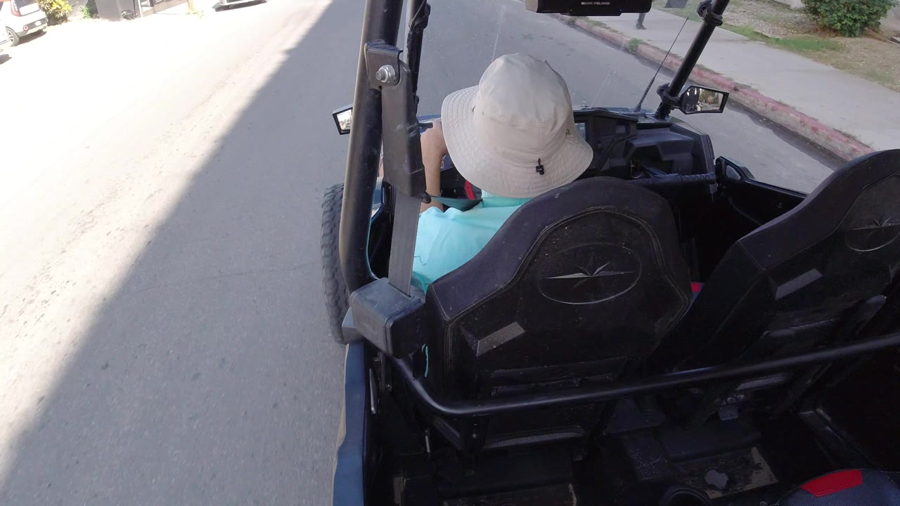 Rear view of a man in a light bucket hat driving a black golf cart along a bright street in Los Cabos, Mexico, with parked cars and building edges ahead.