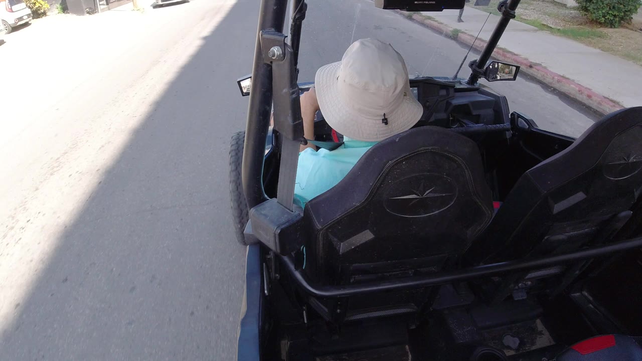 The golf cart moves farther along the Los Cabos street, keeping the driver's hat and seat backs in the foreground while a curb and parked vehicles sit ahead.