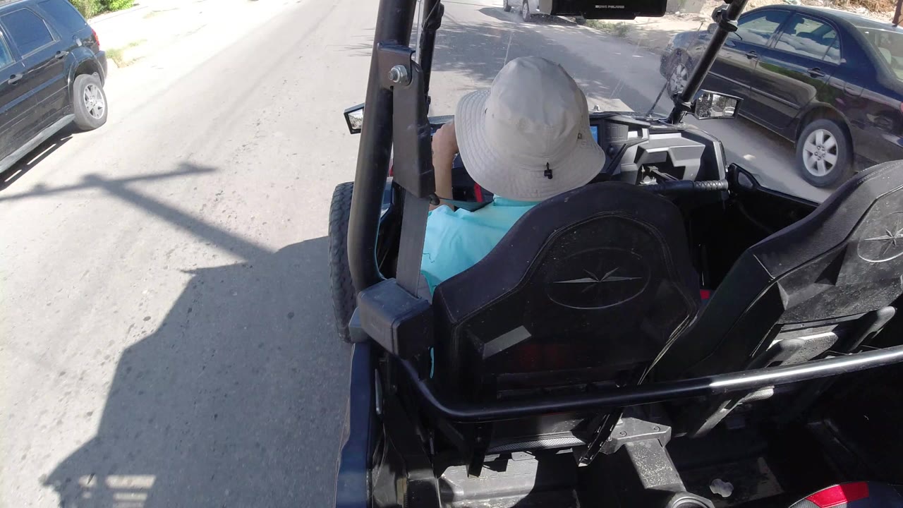 At the start of the Los Cabos clip, a driver in a white sun hat and turquoise shirt sits ahead of the camera in an open black vehicle beside parked cars on a bright road.
