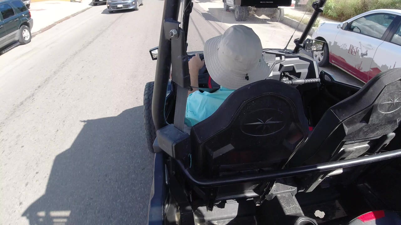 Person in a white hat riding in an open vehicle on a sunny road.