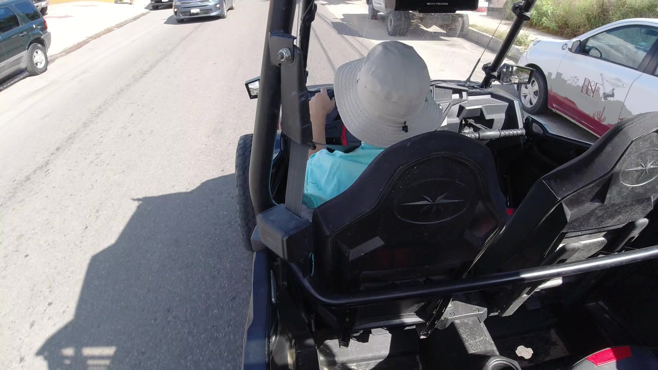 A similar Los Cabos street view shows the white-hatted driver centered ahead, black seat backs in the foreground, and cars parked along the right side of the road.