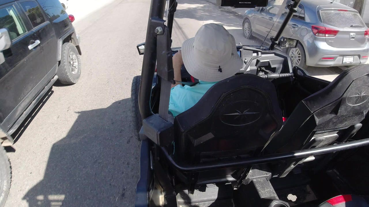 Rear view from an open black vehicle in Los Cabos, Mexico, with a white-hatted passenger seen from behind, parked cars along the street, and no visible faces.
