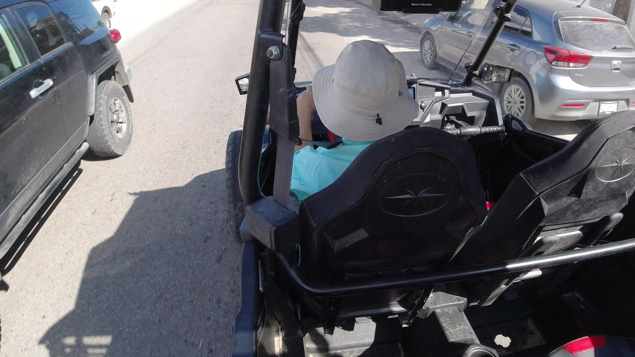 The Los Cabos street view continues from behind the open vehicle, with the passenger's white hat centered and a silver parked car close on the right, with no visible faces.
