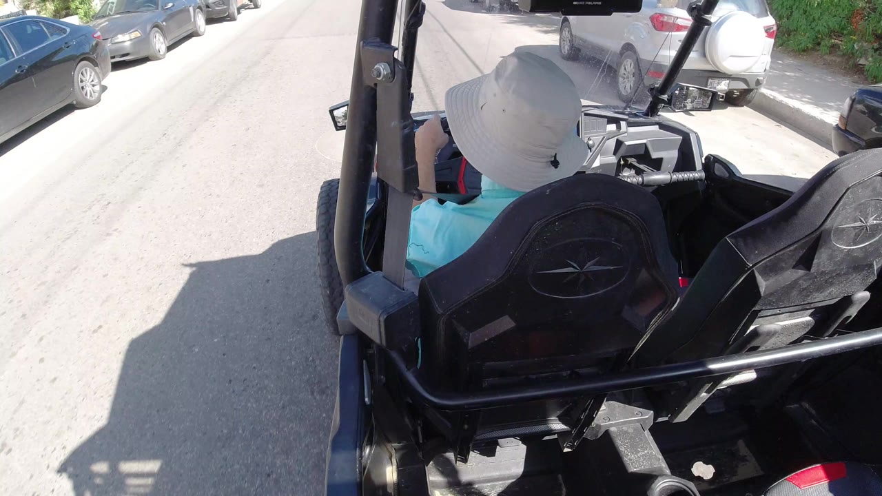 Rear view in Los Cabos, Mexico of a person in a white sun hat and turquoise shirt seated in an open vehicle, with black seats, roll bars, parked cars, and bright road glare visible.