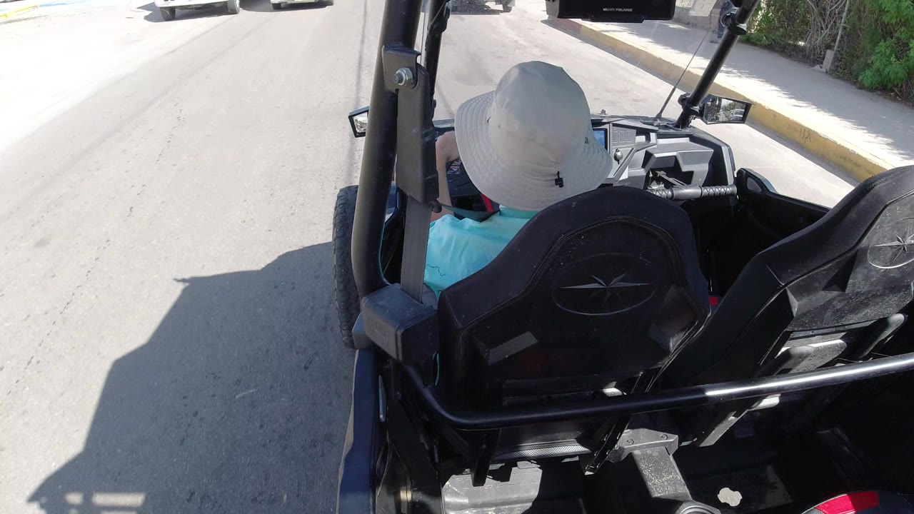A similar forward view from the open vehicle shows the right-side seat, roll bar, pale road surface, curb, and roadside greenery in Los Cabos.