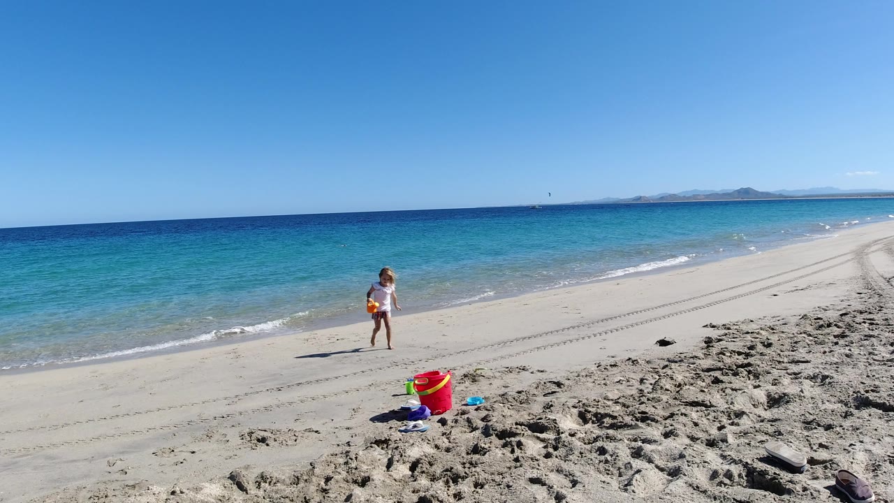 The child stands farther back from the red bucket on the Los Cabos shoreline as small waves break along the beach and the horizon stays clear.