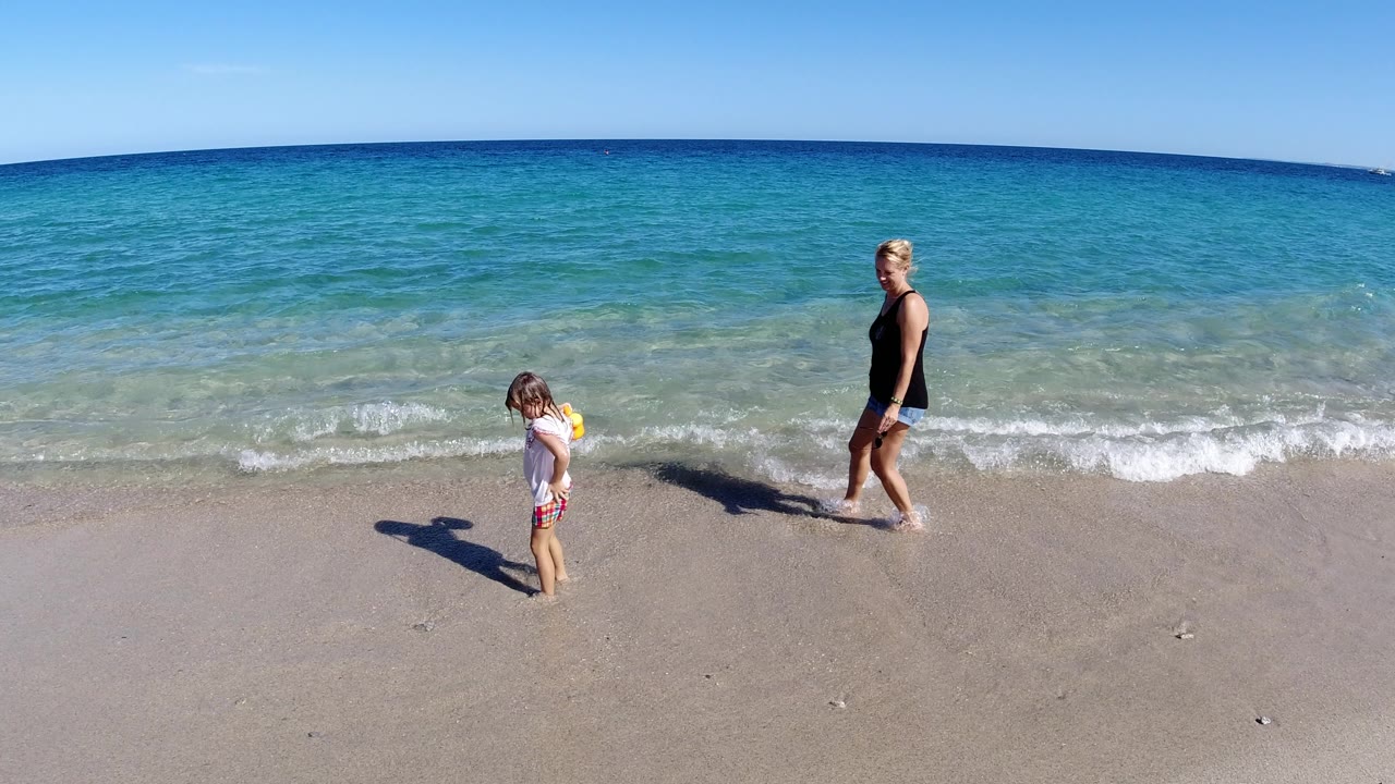 Woman and child walking along a sunny beach beside shallow waves.