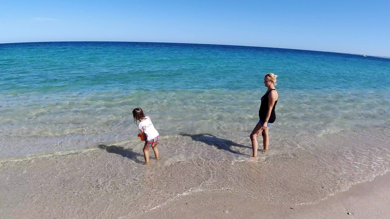 Woman and child standing at the edge of clear water on a sunny beach.