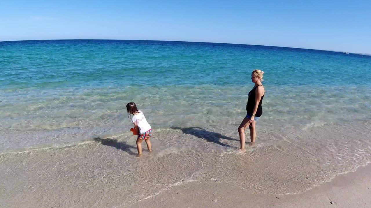 In Los Cabos, Mexico, the child steps closer to the shallow surf while the woman remains upright a few feet back on the beach.