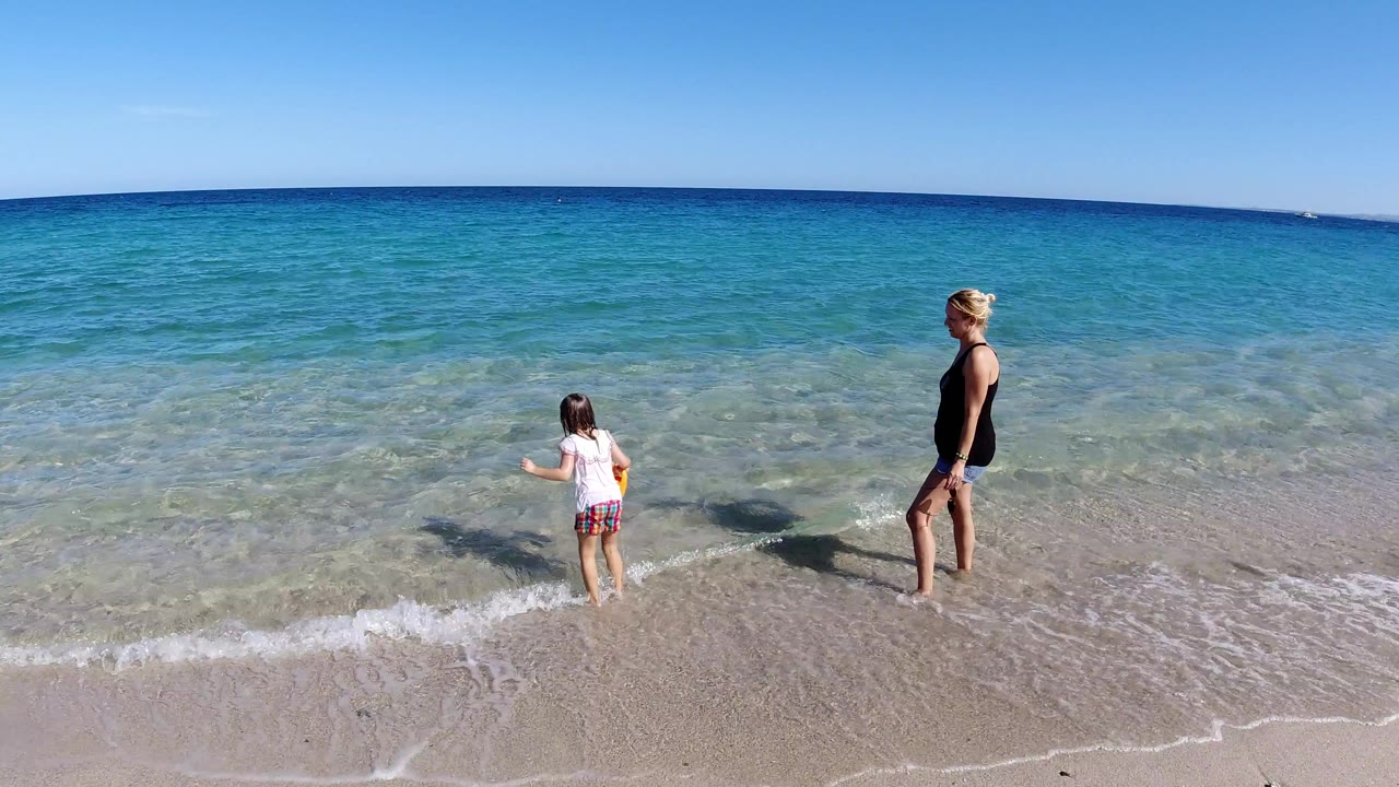 Mother and young daughter standing in shallow surf at a beach.