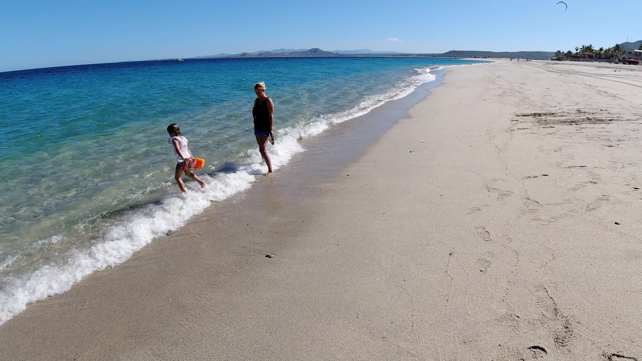 Child carrying an orange bucket near small waves as an adult stands on the beach.