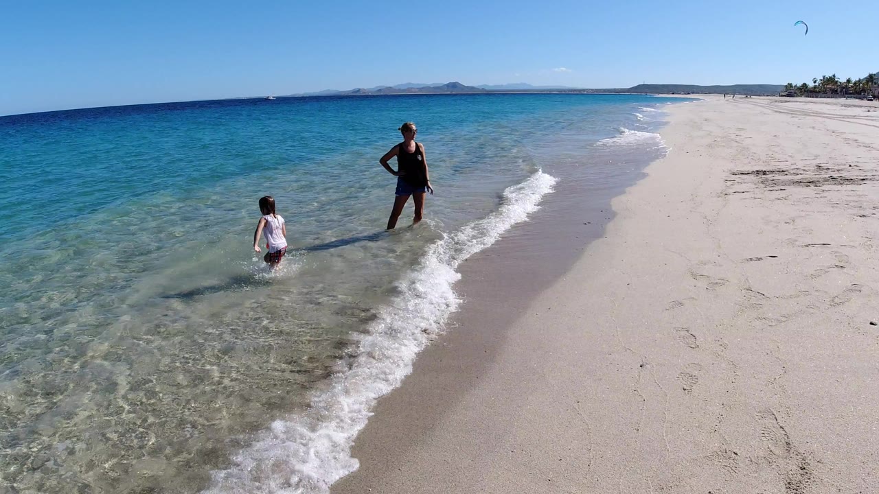 An adult in dark swimwear stands near a child in shallow surf on a Los Cabos, Mexico beach, with clear blue water on the left and pale sand stretching to the right.