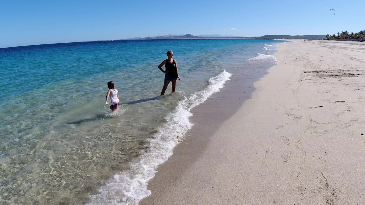 The child stands knee-deep in clear water while the adult faces them from the wet sand, with a narrow white wave curling between them on the Los Cabos shoreline.