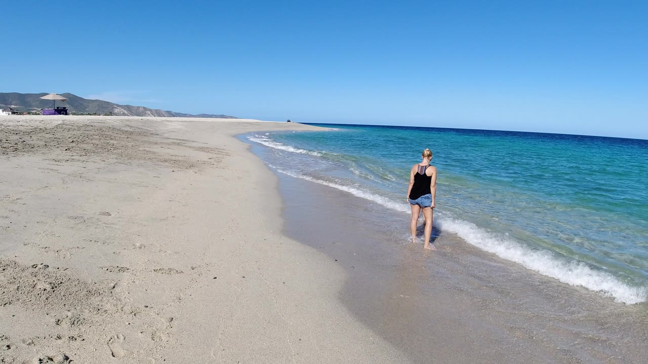 Adult standing at the edge of clear blue surf on a wide sandy beach.