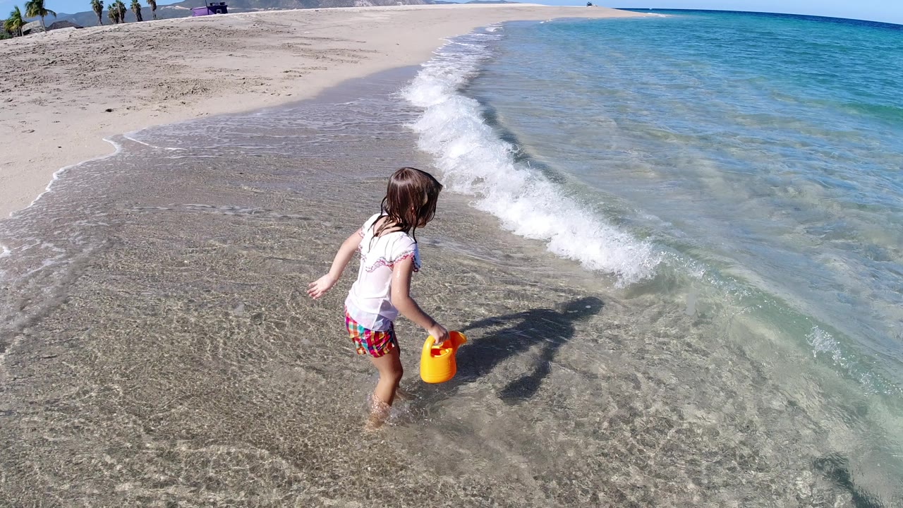 The girl continues along the Los Cabos shoreline with a yellow toy at her side as a small wave curls beside her feet.