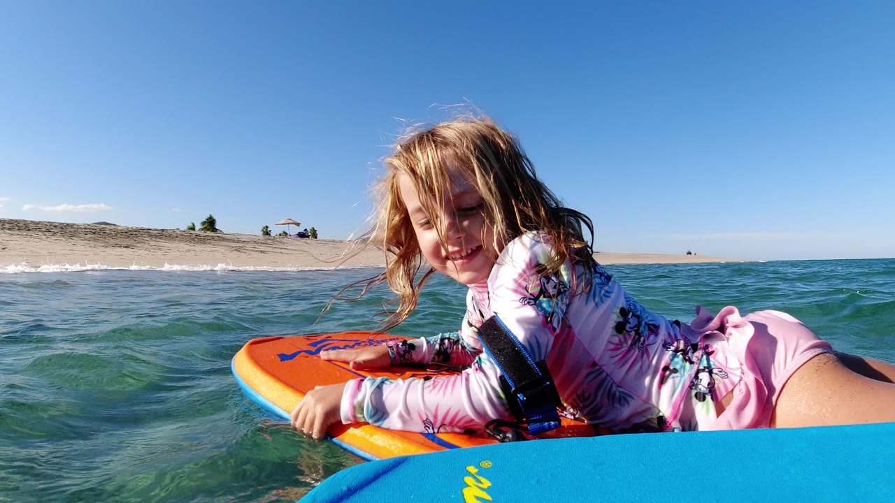 Young girl on an orange boogie board in shallow ocean water near a sandy beach.