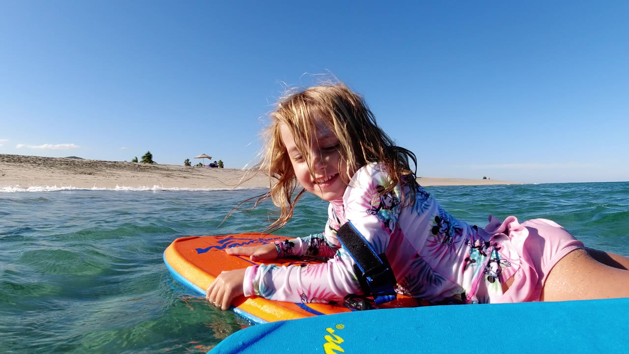 The girl smiles while holding the orange boogie board in Los Cabos, Mexico, with small waves lifting between the camera and the sandy beach.
