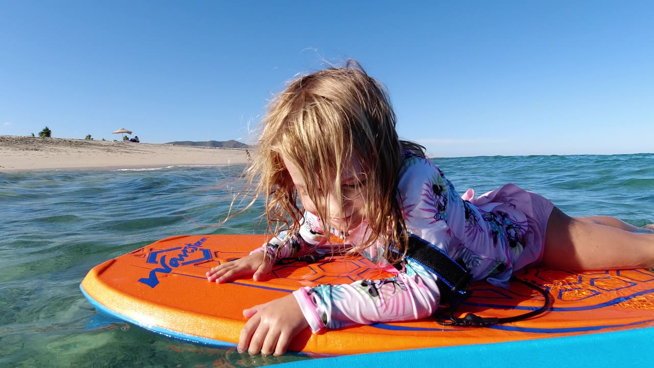 A young child lies face down on an orange flotation board in the ocean at Los Cabos, with wet hair over her face and a sandy beach behind her.