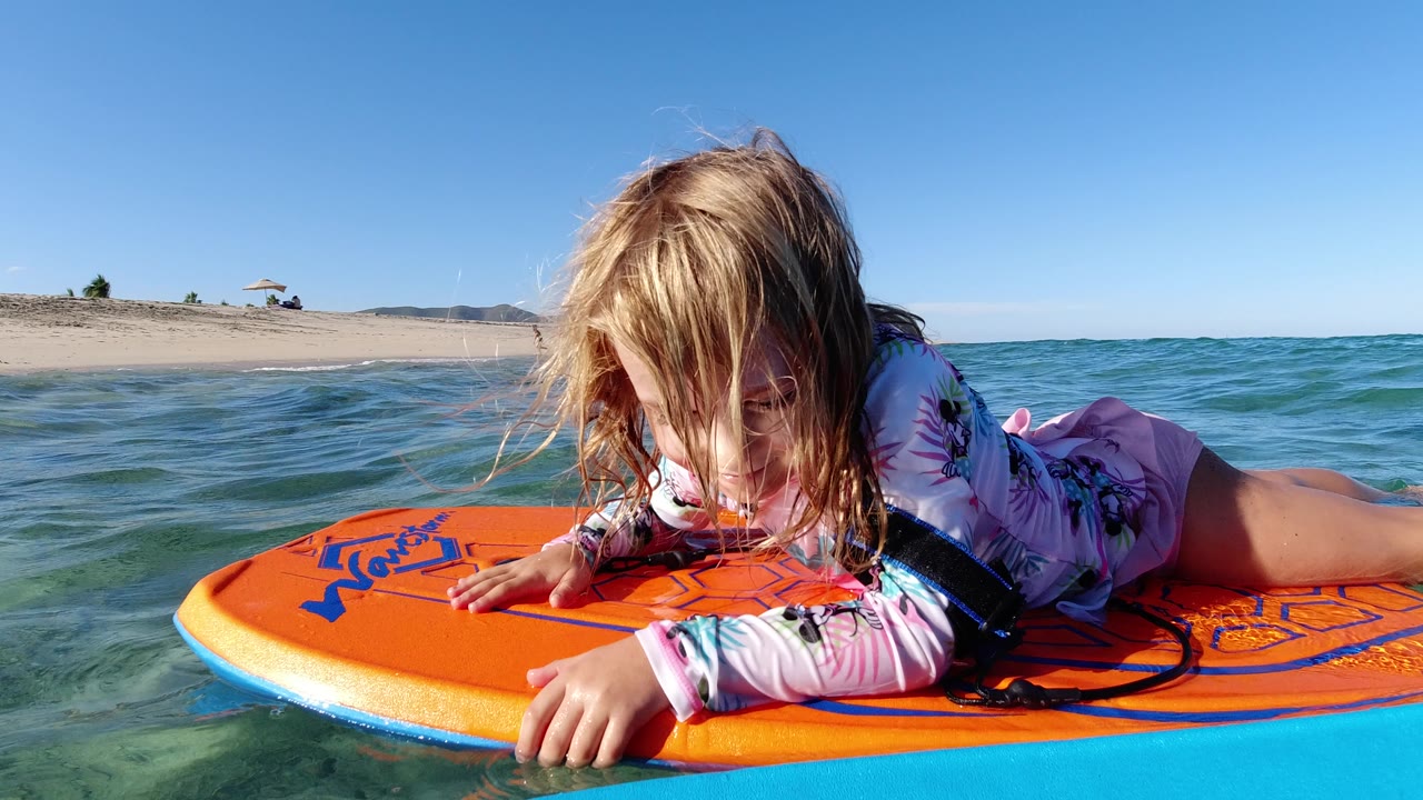The child remains stretched across the orange board in Los Cabos ocean water, with her hand on the front edge and the beach horizon running behind her.
