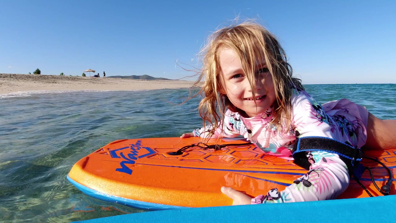 Young blonde girl faces the camera while lying on an orange board in clear shallow water at a Los Cabos, Mexico beach, with sand and blue sky behind her.