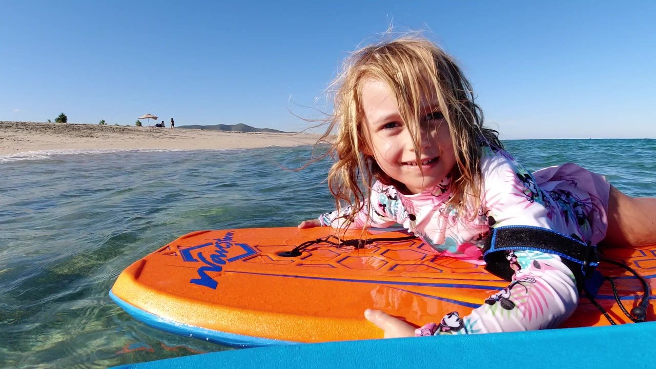 The girl stays close to camera on the orange floating board, with small ripples crossing the clear water and the Los Cabos beach stretching behind her.