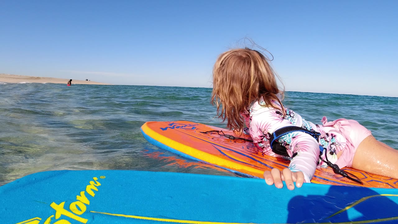 The child remains stretched across the orange board in Los Cabos, Mexico, while the blue board fills the lower foreground and the ocean horizon stays low in the frame.
